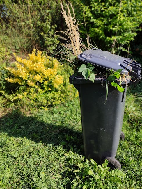 A black wheeled garden waste bin sits on a grassy area in a garden, filled with trimmed garden debris including dried grass, leaves, and small branches. Behind the bin, a vibrant yellow-flowered shrub