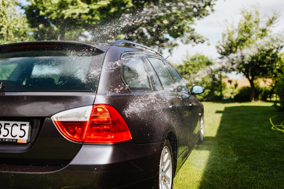 A dark grey hatchback car is being washed outdoors on a sunny day, with water splashing over its rear windshield and body. The car is parked on a well-maintained garden lawn, which features lush green